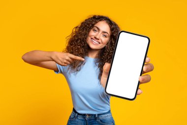Neşeli, kıvırcık saçlı bir kadın boş bir akıllı telefon ekranını işaret ediyor. Uygulama promosyonu için mükemmel, çarpıcı sarı bir arka planı var.