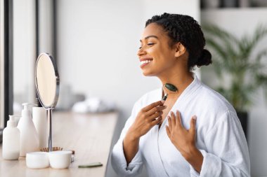 Yeşiltaş Yeşim Roller ile boynuna masaj yapan çekici siyah kadın, gülümseyen Afrika kökenli Amerikalı kadın büyüteç aynasına bakıyor ve cilt bakımı için güzellik aleti kullanıyor.