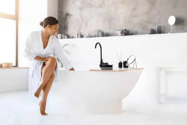 穿着白色浴衣的优雅的黑人妇女坐在现代浴缸旁 美丽的非洲裔美国女士享受着奢华的洗澡时间 用手触摸着水 在时尚的浴室里放松 — 图库照片
