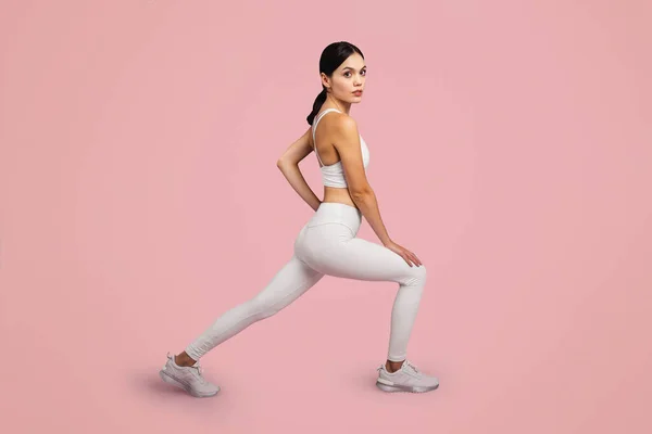 Sporty lady lunging forward exercising during workout over pink studio background, looking at camera, side view. Gym training for muscles flexibility