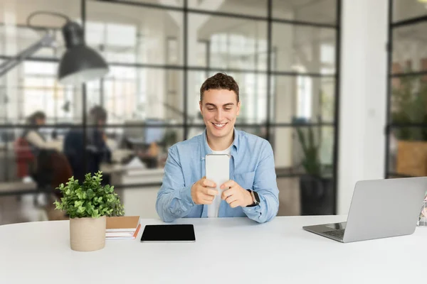 Glad attractive millennial caucasian man manager with laptop, tablet with empty screen at table, chatting at phone in modern coworking office. Work, business app, professional career