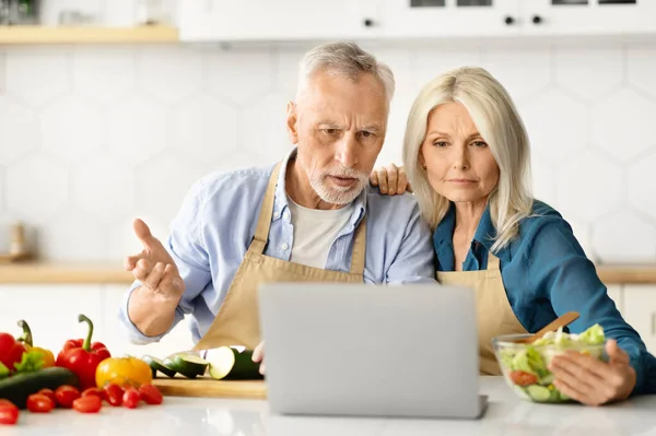 Wrong Recipe. Confused Senior Spouses Looking At Laptop While Cooking In Kitchen, Upset Elderly Man And Woman Having Problem With Computer, Reading Bad News While Preparing Food At Home