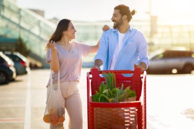Gülümseyen genç orta doğu çifti güneşli günün tadını çıkarırken park yerinde içi taze yiyeceklerle dolu kırmızı alışveriş arabalarını itiyor.
