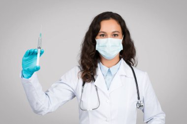 Beyaz önlüklü, koruyucu maskeli ve eldivenli kendine güvenen kadın doktor gri stüdyo arka planında izole edilmiş şırınga gösteriyor. Hastalık tedavisi, aşı, ilaç ve sağlık hizmetleri