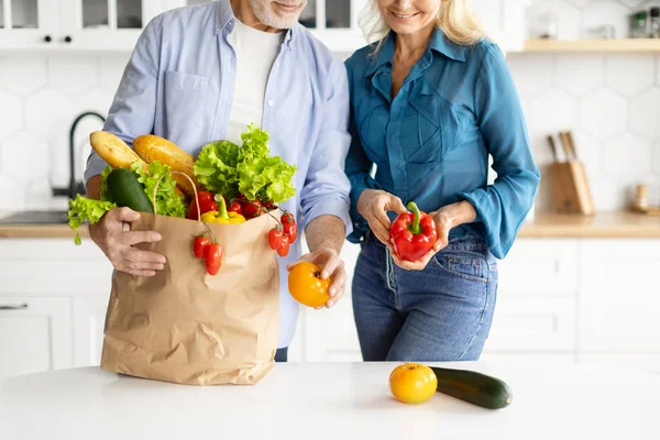 Smiling elderly couple unpacking bag of fresh, colorful vegetables and bread in their bright modern kitchen, happy senior spouses return to home after food shopping, checking groceries, cropped shot