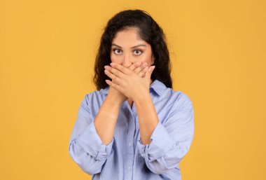 Koyu kıvırcık saçlı endişeli genç bir kadın elleriyle ağzını kapatıyor, gözleri endişelerini iletiyor ya da sessiz kalma arzusunu dile getiriyor, düz sarı bir arka plana karşı.