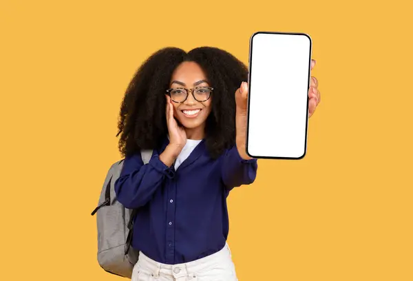明るい黄色の背景に灰色のバックパックと アプリのプレゼンテーションに理想的な空白のスマートフォン画面を表示するメガネの喜ばしい若い女性 — ストック写真