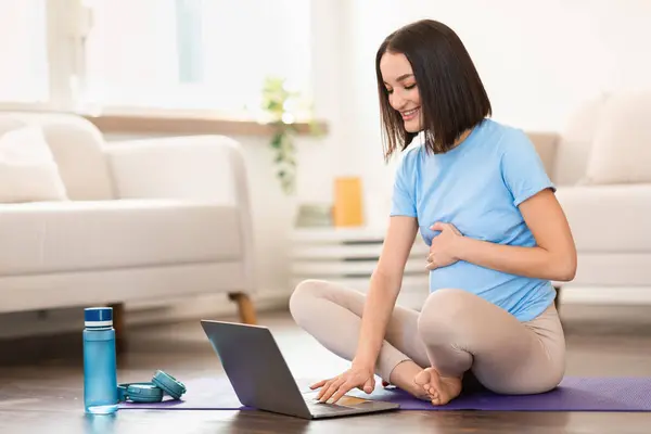 Happy young pregnant woman watching video workout lesson on laptop, sitting on mat near computer ready for prenatal yoga class, training in living room interior. Motherhood, pregnancy
