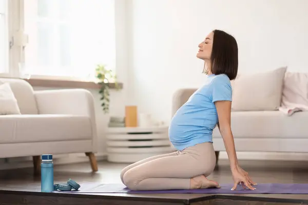 年轻孕妇在室内冥想或瑜伽锻炼期间做背弯的侧视图 穿着休闲裤的准妈妈在室内垫子上做呼吸练习 — 图库照片