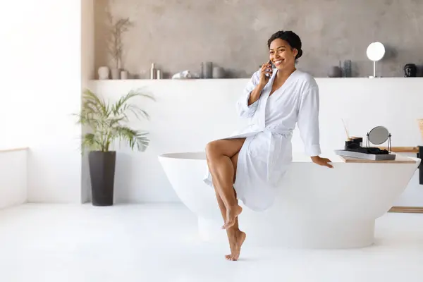 現代の浴室で携帯電話で話すバスローブを身に着けている美しい黒人女性 笑顔のアフリカ系アメリカ人の女性はバスタブに横たわり 家で携帯電話で楽しい会話をする コピースペース — ストック写真