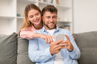 Milenyum çifti akıllı telefonlarına dalıp sosyal medyaya ve mesajlara bakıyorlar, rahat ev ortamında modern iletişimi örnek gösteriyorlar, oturma odasındaki kanepelerinde oturuyorlar.