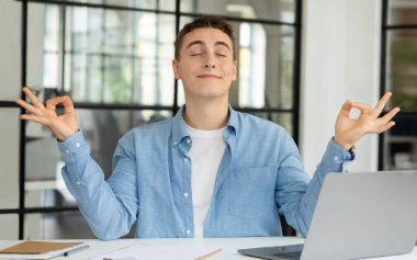Masasında dizüstü bilgisayarı olan, meditasyon yapan, modern çalışma ofisinde nefes egzersizlerinin keyfini çıkaran, gülen bin yıllık beyaz adam yöneticisi. İş, iş molası, başarı ve boş vakitlerde rahatlama.