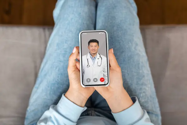 Virtual Medical Consultation. Sick Woman Talking With Asian Male Doctor Online Via Video Call On Cellphone While Sitting On Couch At Home, Therapist Consulting Patient From Smartphone Screen, POV