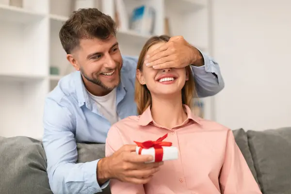 Romantic Present. Young Couple Celebrating Valentines Day Holiday, Husband Congratulating Wife Giving Gift Box And Closing Her Eyes At Home. Birthday And Anniversary, Family Holiday