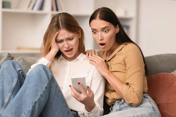 Unhappy young woman shares shocking text message on smartphone with friend, sitting on sofa indoors. revealing her partners infidelity, reflecting betrayal and disbelief.