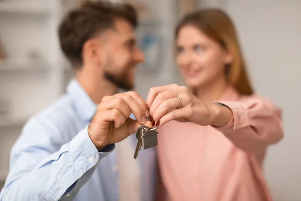 Real estate offer. Millennial European couple hugging showing keys to new apartment indoor. Focus on key to first own house. Smiling millennial man and woman celebrating relocation moving new home