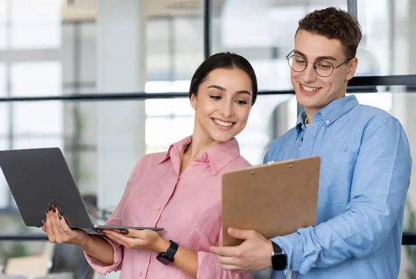Glad millennial european man show clipboard to arab lady manager, planning project, startup together, in modern coworking office. Professional work and business meeting, idea creation