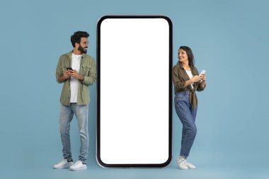 Tarz sahibi genç Hintli erkek ve kadın telefonları kullanıyor ve beyaz ekranlı büyük akıllı telefonlara bakıyor, en yeni mobil uygulamaları kontrol ediyor, mavi stüdyo arka planında izole edilmiş, model, boş alan