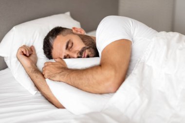 Adam rahat bir şekilde battaniyeye sarılı olarak yatakta uyuyor, dinlendirici bir uykuda beyaz yastığa sarılıyor ve modern yatak odası ortamında dinleniyor. Kestirirken ve eğlenirken çekilmiş bir fotoğraf.