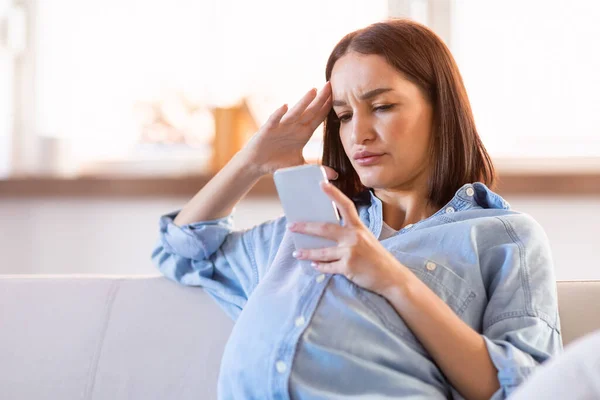 Doomscrolling. Discontented Pregnant Young Lady Websurfing On Cell Phone Sitting On Couch At Home. Stress Of Social Networking and Online Communication Problems in Pregnancy
