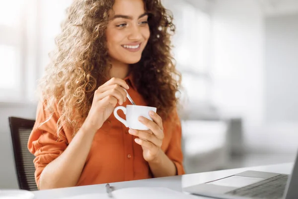 Smiling curly-haired professional enjoying a warm cup of coffee during a break, with a laptop on the desk in a bright office setting, free space