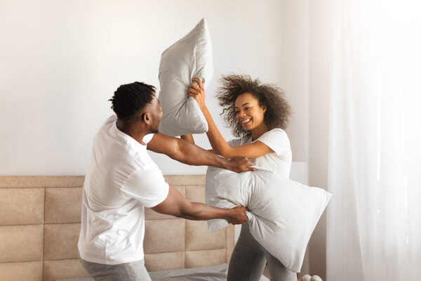 Joyful African American Millennial Couple Having Pillow Fight For Fun In Bedroom At Home. Young Husband And Wife Fighting With Pillows, Laughing And Flirting On Weekend. Happy Romantic Relationship