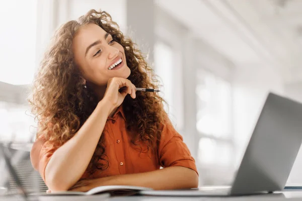 Cheerful curly-haired professional enjoying a creative moment, holding pen to chin and smiling at laptop in a modern, sunlit office space
