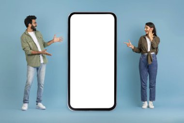 Şaşırtıcı duygusal milenyum Hintli erkek ve kadın aşıkları beyaz ekranlı büyük bir telefonu işaret ediyor, harika mobil uygulama, online teklif, mavi stüdyo geçmişi, model, boş alan