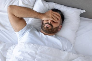 Uykulu ve stresli bir adam yatakta, yüzü elle örtülmüş, akşamdan kalma belirtileriyle boğuşuyor ya da yatak odasında ateşi var. Kronik baş ağrısı ve kapalı yerde yatan uykusuzluk çeken bir adam.