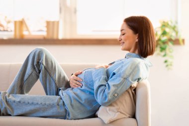 Neşeli hamile kadın koltukta dinleniyor ve anneliği bekliyor, elleri karnının üstünde dinleniyor, doğmamış çocuğuyla olan bağını simgeliyor. Yan görüntü. Hamilelik mutluluğu