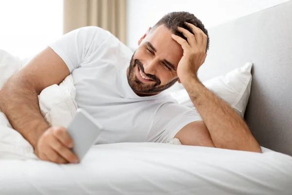 Smiling bearded caucasian man lying in bed and using cellphone, sending messages and emails in morning at cozy bedroom interior. Guy websurfing via phone and scrolling social media after sleep