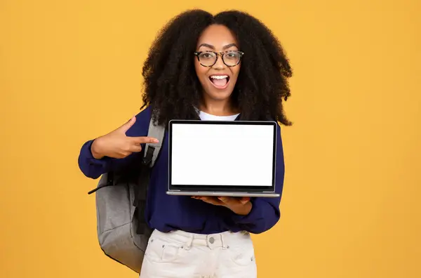 Enthusiastic young black woman with glasses pointing to blank laptop screen, perfect for mockups, with backpack, against yellow background