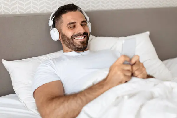Middle aged European man lying in bed with earphones and phone, listens to audiobook or music online on his smartphone, resting in modern bedroom. Morning relaxation leisure