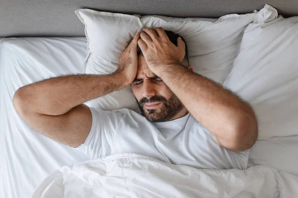 Bearded European man touching head with pained expression, suffering from headache, lying in bed in his bedroom. Healthcare and sickness, painful migraine issue concept