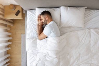 Sakallı bir adamın modern yatak odasında rahat bir şekilde uyuduğu beyaz çarşaflar, yastıklar ve battaniyelerle rahat bir yatakta uyuduğu görülebilir. Huzurlu dinlenme, kaliteli uyku konsepti