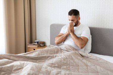 Hasta adam kağıt mendile burun sümkürerek soğuk algınlığı ve grip belirtileri gösteriyor evdeki modern yatak odasında battaniyeyle kaplı olarak yatakta oturuyor. Sinüzitten muzdarip bir adam kendini iyi hissetmiyor.