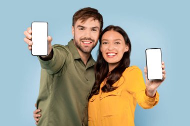 Neşeli genç adam ve kadın akıllı telefonlarını boş ekranlarla sunuyorlar. Model görüntüler için ideal, yumuşak mavi bir arka plan,