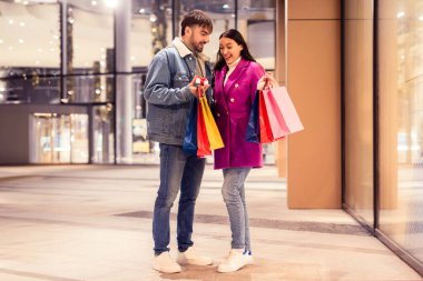 Millennial koca karısını küçük bir paketle şaşırtır, alışveriş merkezinin dışında elinde poşetlerle gülümseyerek sunar. Romantik hediye, kış akşamı konsepti.