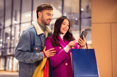 Birçok alışveriş çantası olan mutlu genç müşteriler akıllı telefonlara bakıyorlar, kış tatili alışverişleri ve satışları arıyorlar, modern alışveriş merkezinin önünde birlikte duruyorlar. Noel çılgınlığı