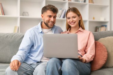 Mutlu genç çift dizüstü bilgisayarlarıyla kanepede oturuyor, evde online olarak eğleniyorlar. Neşeli eşler internette geziniyor ve hafta sonları film seyrediyor.