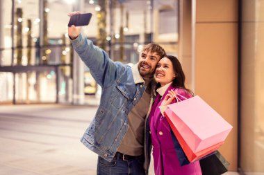 Milenyum çifti alışveriş merkezinin dışındaki akıllı telefondan selfie çekiyor, alışveriş torbalarıyla poz veriyor, kış akşamları cep telefonu uygulaması kullanarak alışveriş çılgınlıklarını yakalıyor..