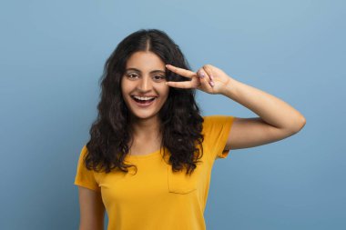 Sarı tişört giyen neşeli, genç Paskalya 'lı bayan öğrenci gözlerinin yanında V işareti gösteriyor ve kameraya gülümsüyor, mavi stüdyo arka planında izole bir şekilde poz veriyor. Gen Z, yakınlaştırıcı konsepti
