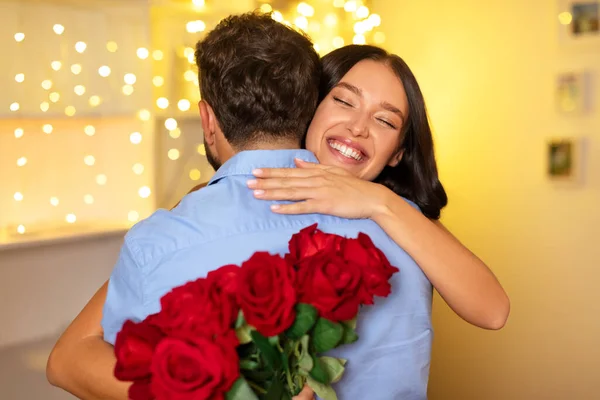 女人带着灿烂的笑容 从背后拥抱着男人 手里拿着一束美丽的红玫瑰 背后点着温暖的吊灯 — 图库照片