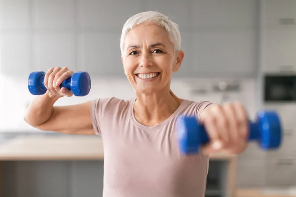 Home workout. Happy mature woman exercising with dumbbells smiling to camera indoors, portrait of healthy fitness lady training in domestic interior, keeping fit, leading active sporty lifestyle