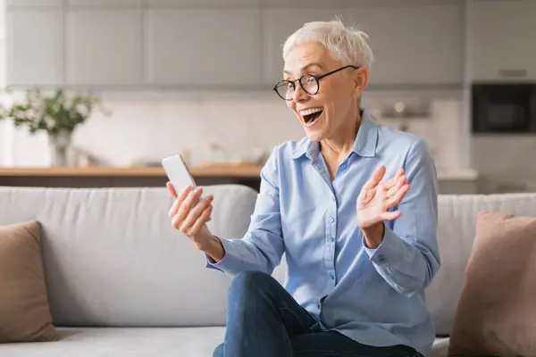 Excited emotional mature lady holding smartphone reading wow online offer, sitting on couch at modern living room interior. Woman with gray hair looking at phone gadget in excitement, uses new app