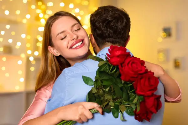 背後から男を抱きしめる明るい笑顔の女性 赤いバラの花束をクラッチ 居心地の良い背景を提供するツインリング文字列ライト — ストック写真