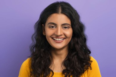 Mor renkli stüdyo arka planında izole edilmiş sarı tişört giyen, kameraya gülümseyen neşeli, kıvırcık, genç Hintli kız öğrenci. Gen Z, yakınlaştırıcı konsepti