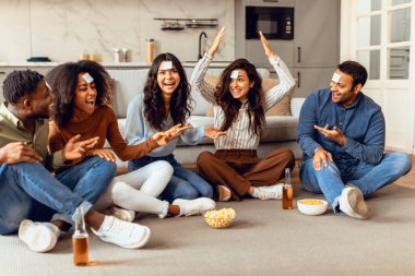 Neşeli çok kültürlü öğrenciler, tahmin edin kim oyun oynuyor, bira ve patlamış mısırdan hoşlanıyor, modern oturma odasında yerde oturuyor, hafta sonları evde birlikte takılıyor, alnında kartlar olan