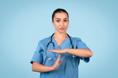Mavi önlüklü, kendine güvenen genç hemşire el hareketi yapıyor, muhtemelen bir mola veriyor ya da klinik bir ortamda duraklama ihtiyacı duyuyor, mavi arka plan.
