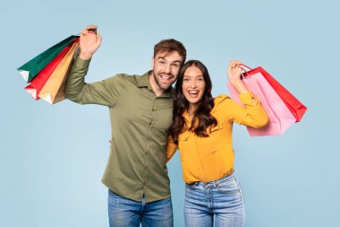 Genç adam ve kadın neşeyle renkli alışveriş torbalarını gösteriyor, mavi bir arka planda cümbüşlerinden mutluluk ve memnuniyet yayıyorlar.